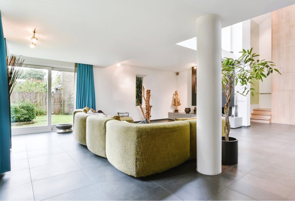 Modern living room with green curved sofas, large windows with blue curtains, indoor plants, light wood stairs, and a view of a garden outside.