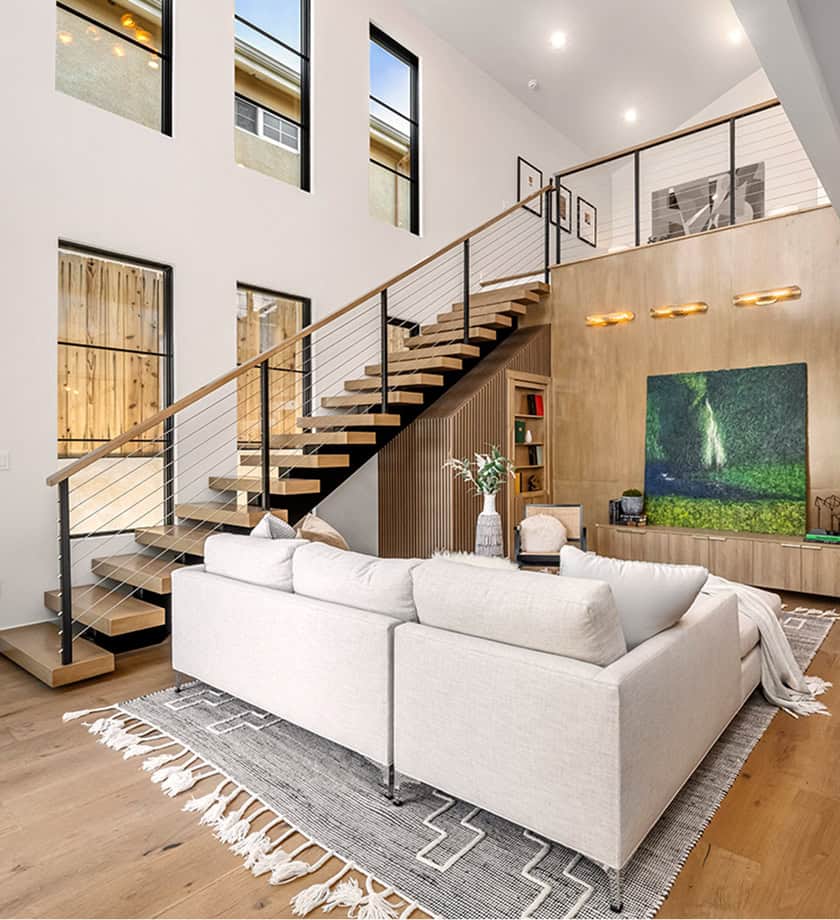 Modern living room with a white sectional sofa, light wood floors, open staircase, large windows, and a wall-mounted TV below a loft area.