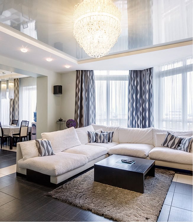 Modern living room with a large white sectional sofa, patterned curtains, a rectangular coffee table, a rug, and a chandelier, with dining area visible in the background.