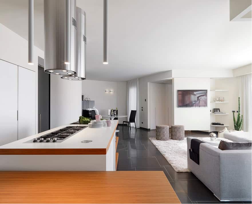 Modern open-plan kitchen and living room with a kitchen island, bar stools, gray sofa, wall-mounted TV, and large windows letting in natural light.